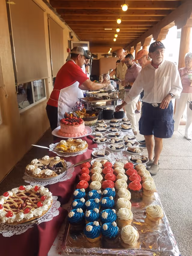 A long buffet table of cupcakes, pies and a cake on a covered outdoor patio while staff serve residents.