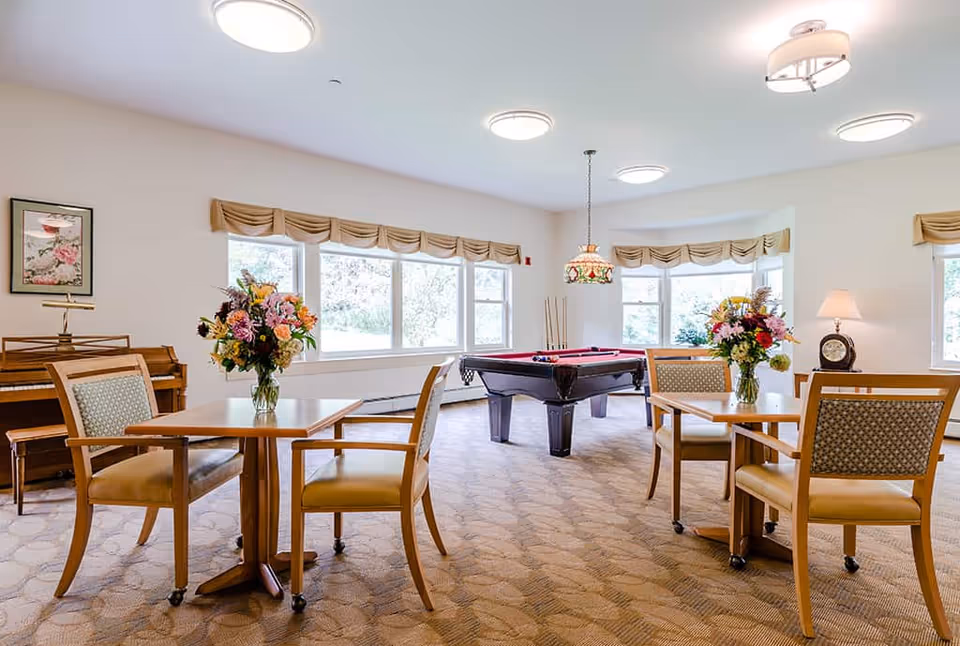 A bright and spacious common room with large windows letting in natural light. The room features two wooden tables each with four cushioned chairs and a vase of colorful flowers on top. In the center, there is a pool table with a red felt surface and pool cues resting on it. A piano is visible against the wall on the left side, and a table lamp and clock are on a small table near the windows on the right. The room has beige carpet and light-colored walls with valance curtains on the windows.