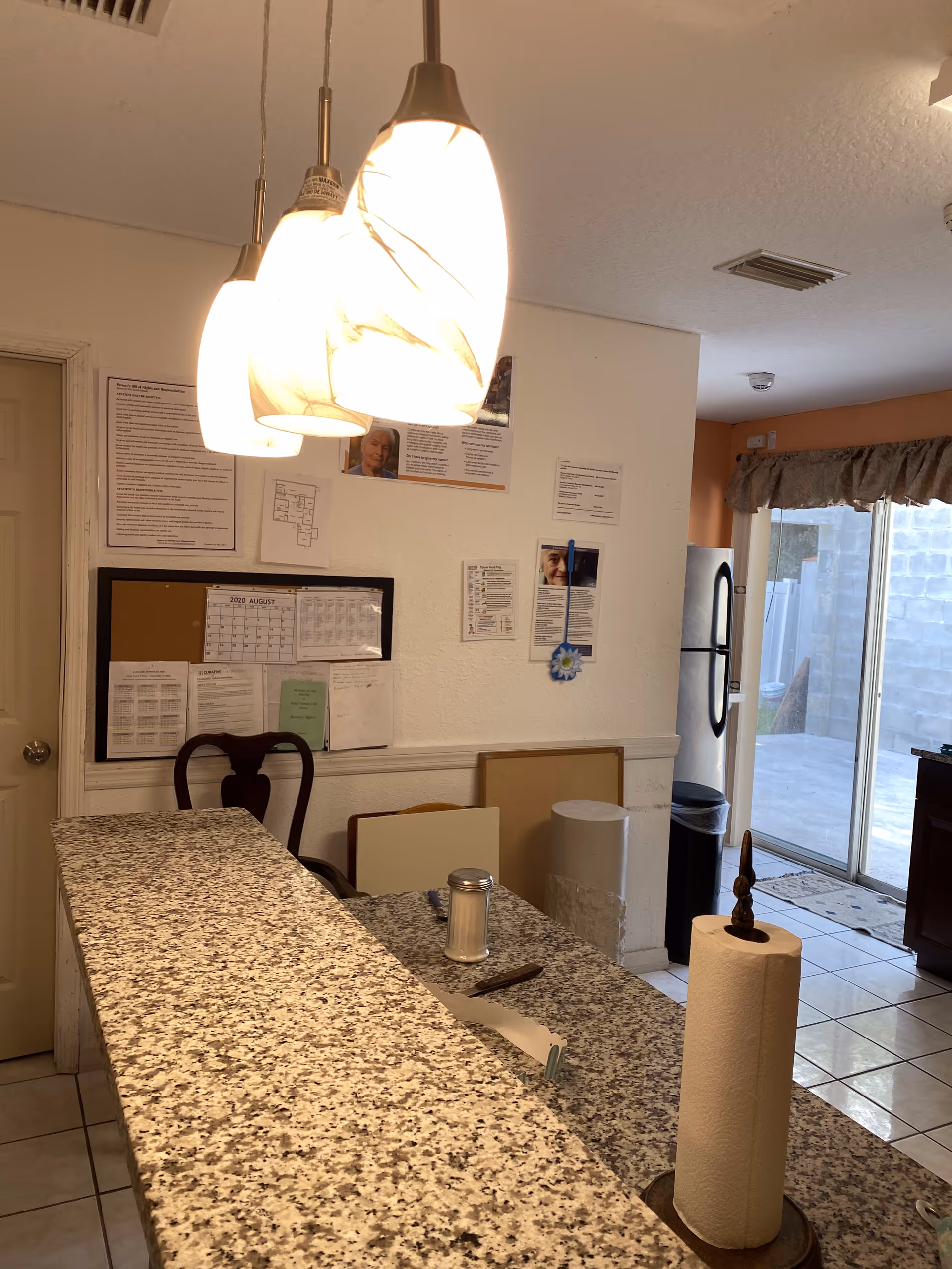 Granite countertop kitchen island with pendant lights, a paper towel holder and notes on a wall near a refrigerator and sliding glass door.
