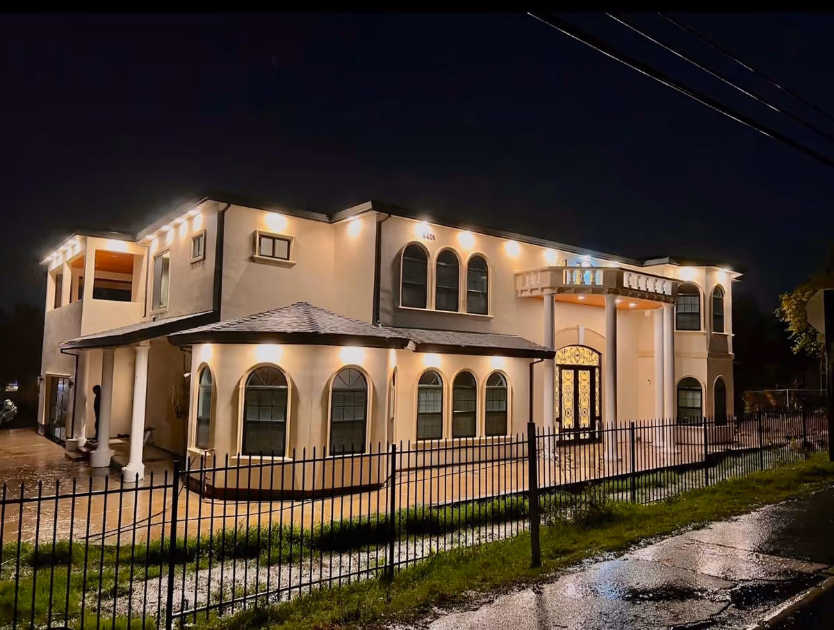 Night view of a two-story building with exterior lights illuminating the facade. The building has arched windows, a balcony with columns, and a decorative front door. A black metal fence surrounds the property, and the ground appears wet from rain.