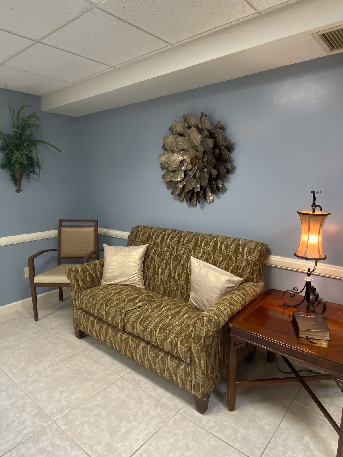 A small sitting area with a patterned brown loveseat with two light-colored pillows, a wooden side table with a decorative lamp and a book titled 'Holy Bible', a wooden chair with beige upholstery, a wall-mounted green plant, and a large metallic flower wall decoration on a light blue wall.
