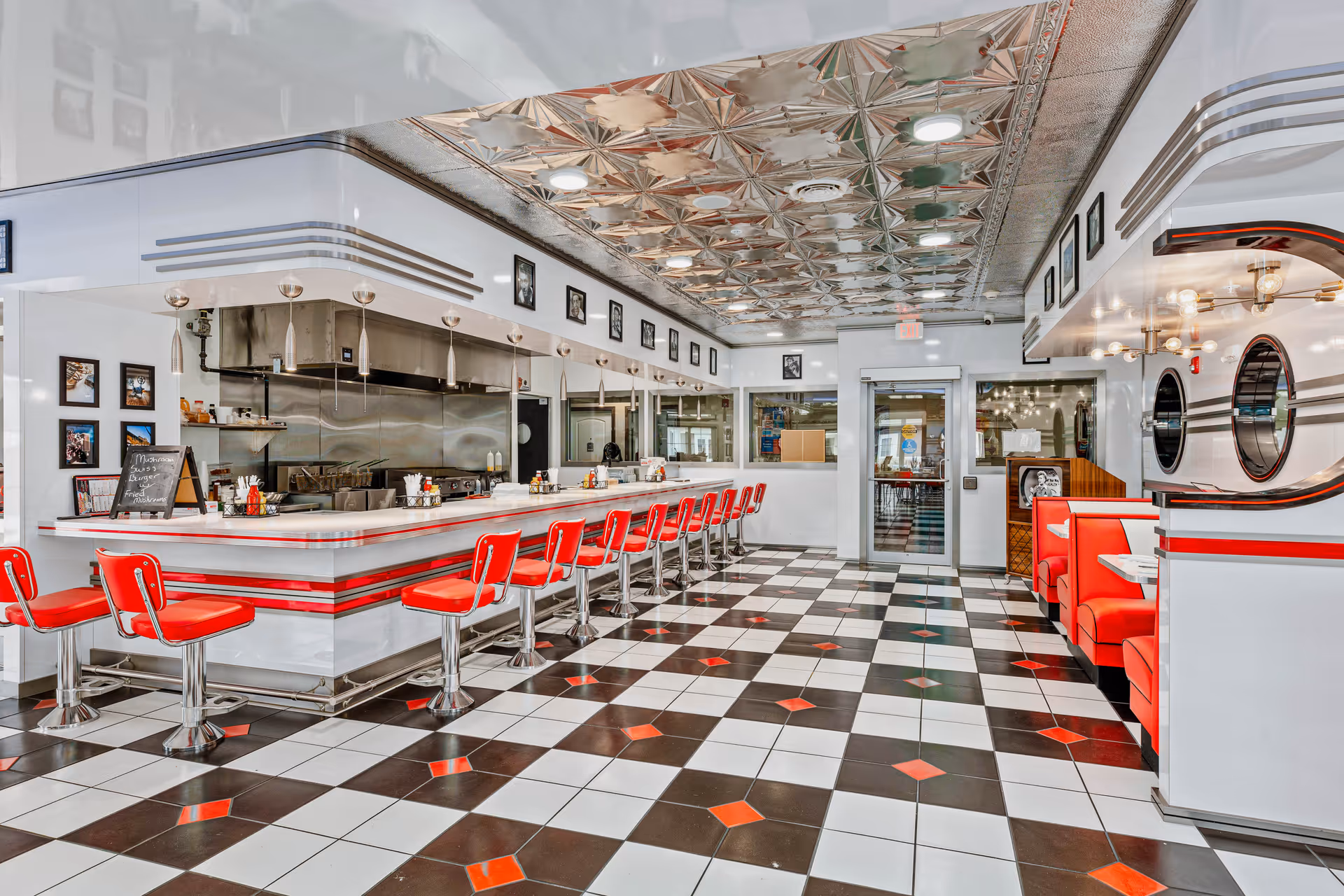 Interior view of a retro-style diner with a long white counter and red bar stools. The floor has a black and white checkered pattern with red diamond accents. There are red booth seats on the right side and a stainless steel kitchen area behind the counter. The ceiling has a decorative metallic pattern with recessed lighting.