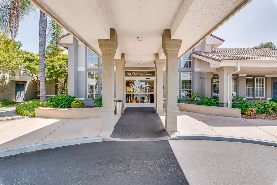 Entrance of a senior living facility with a covered driveway supported by columns, landscaped greenery on both sides, and glass doors with a 'Welcome Home' sign above them.