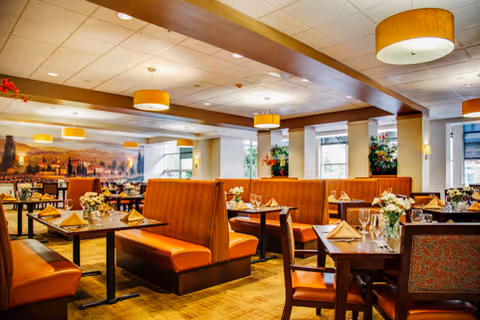 Bright, warmly lit dining room with orange-upholstered booths and tables set with napkins and floral centerpieces.