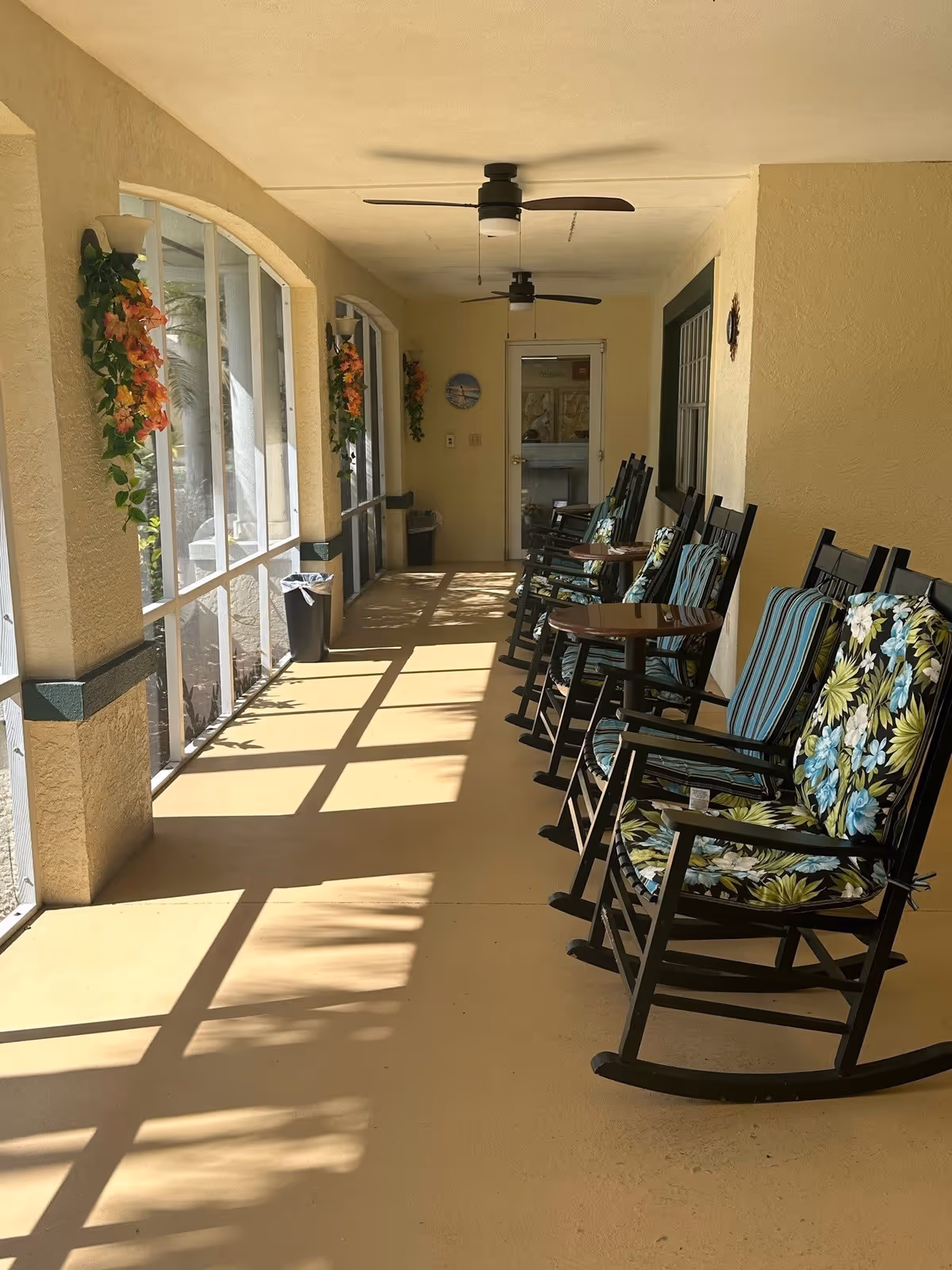A sunlit screened porch with several black rocking chairs featuring floral and striped cushions lined up along the right side. Small round tables are placed between some of the chairs. The left side has large screened windows with hanging flower decorations. Two ceiling fans are mounted on the porch ceiling, and a door is visible at the far end.