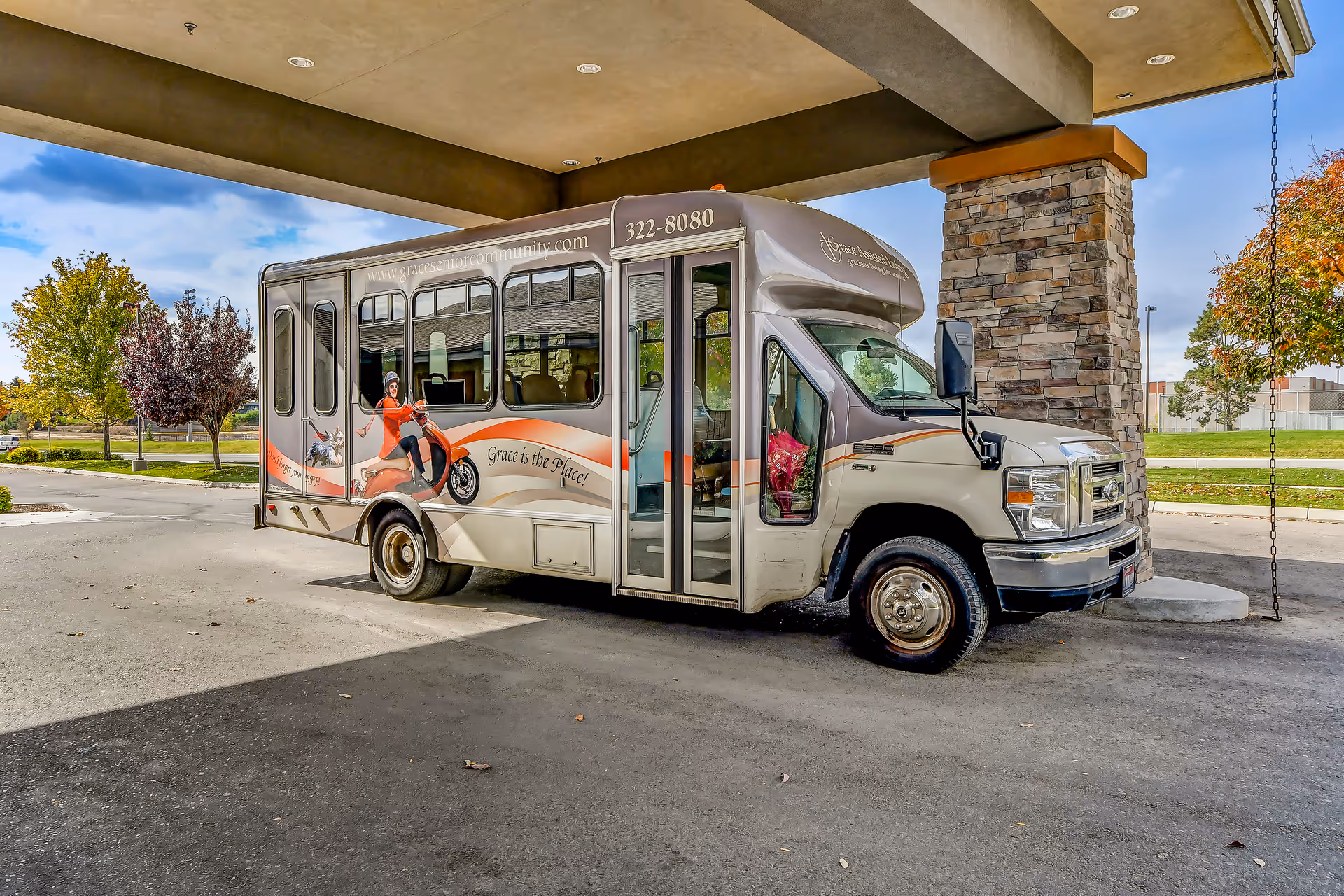 A beige and orange shuttle bus parked under a covered driveway with stone pillars. The bus has the text 'Grace is the Place!' and 'www.graceseniorcommunity.com' on its side, along with an image of a woman riding a scooter. Trees with autumn foliage and a grassy area are visible in the background.