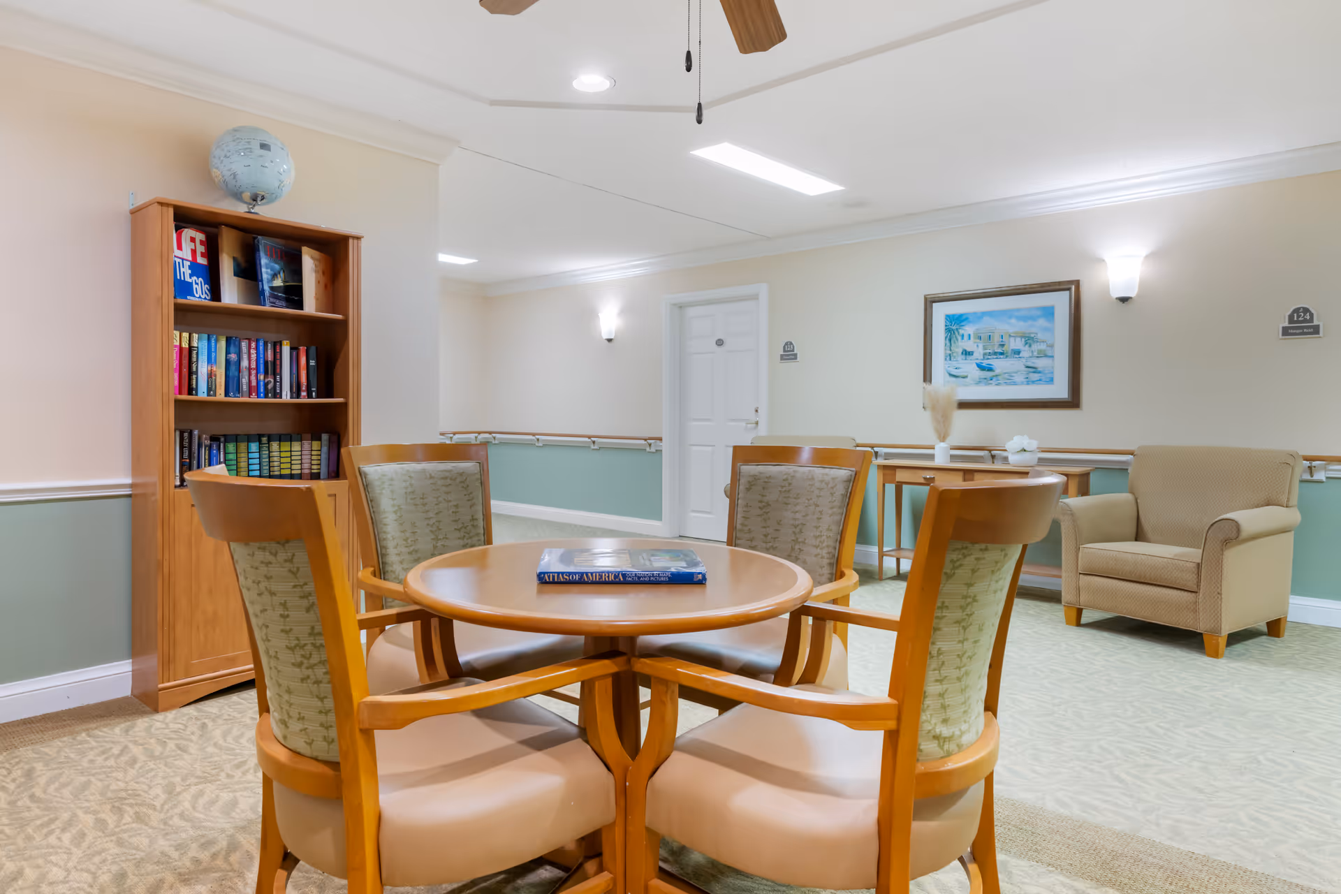 A bright communal seating area with a round wooden table and four chairs, a bookshelf, armchair, and framed artwork in a senior living facility.