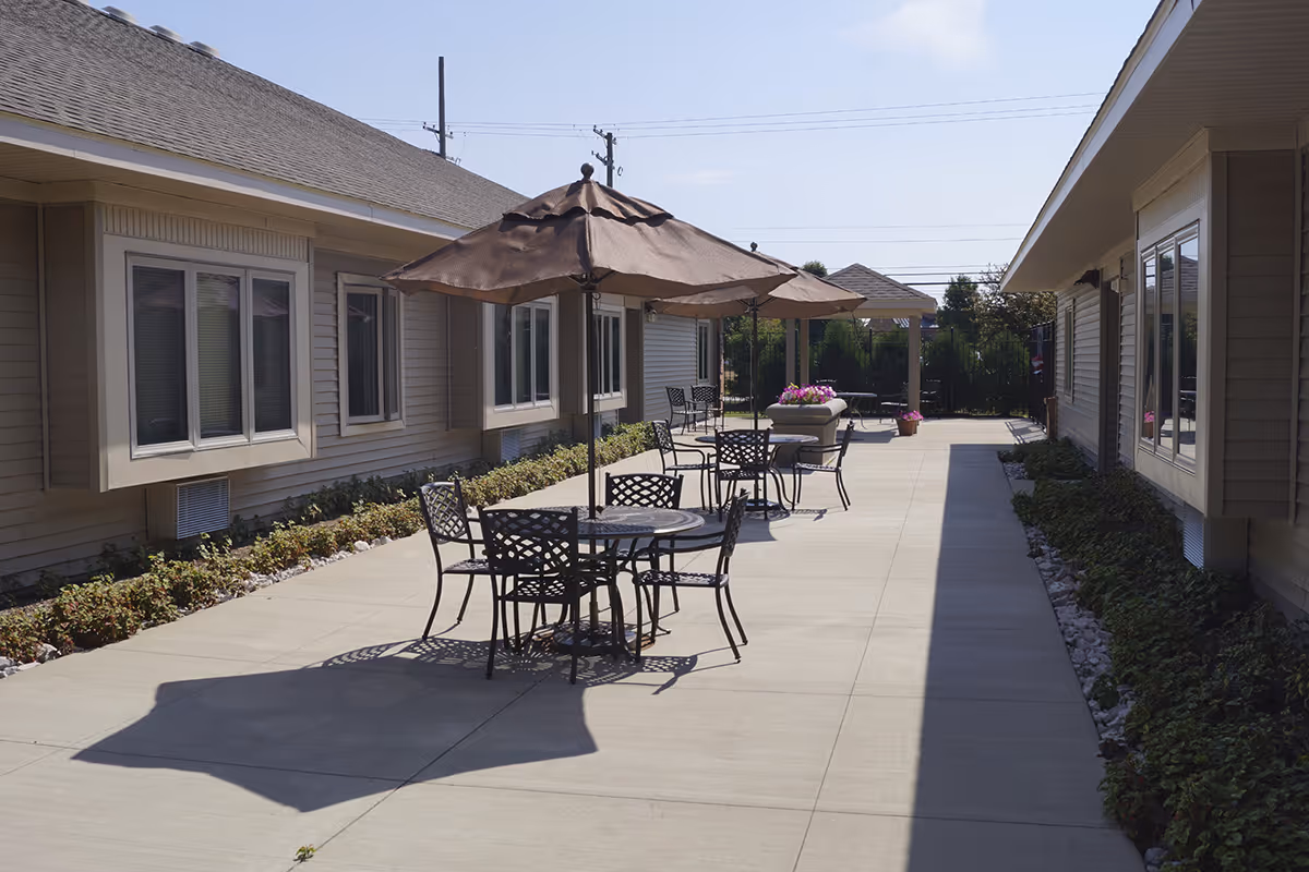 Outdoor patio area between two buildings with several round metal tables and chairs, each table shaded by a large brown umbrella. There are plants and shrubs along the sides of the buildings, and a clear blue sky overhead.