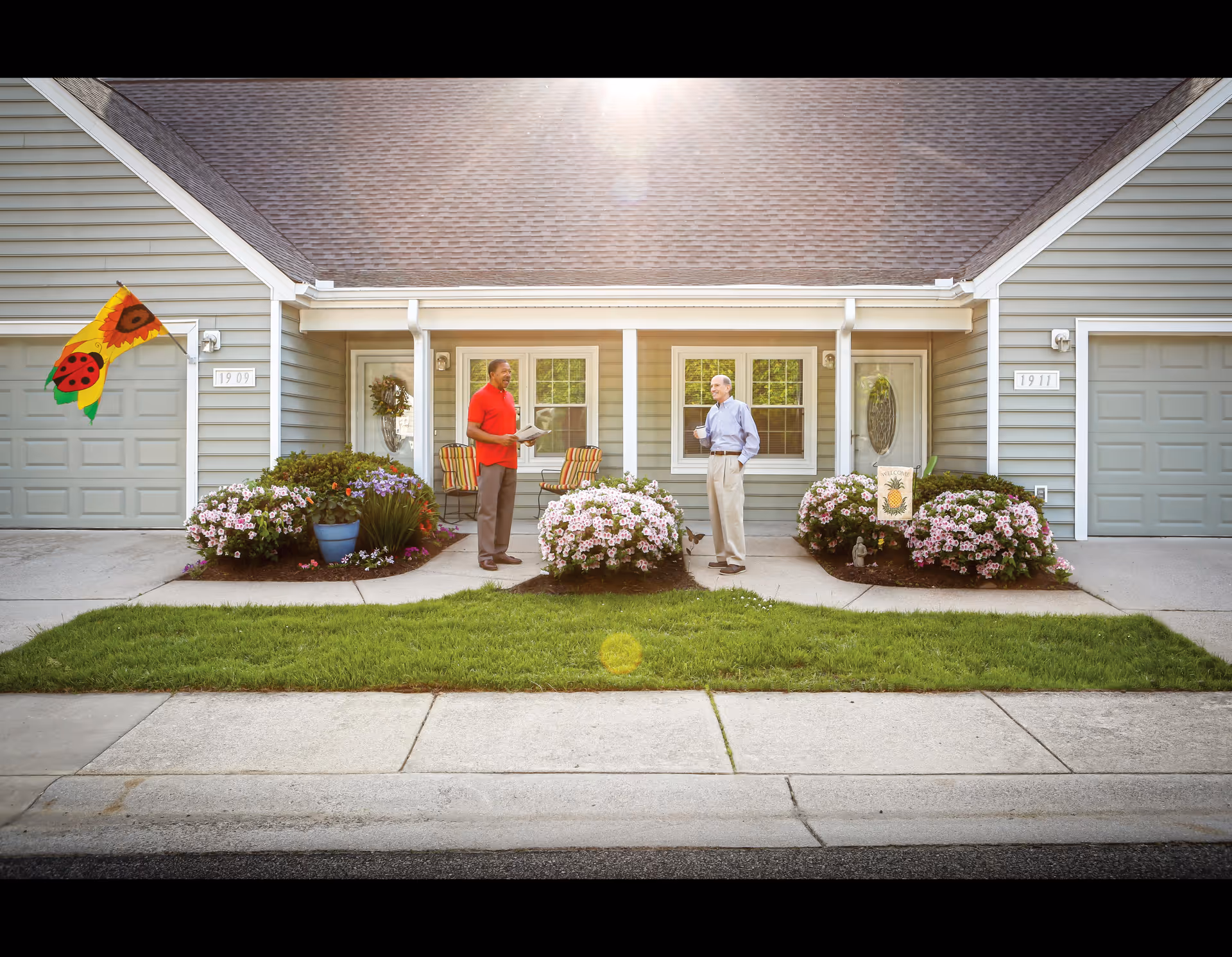 Two elderly men standing outside the front doors of adjacent units in a retirement community, engaged in conversation. The units have light gray siding, white trim, and attached garages. There are flower beds with blooming pink flowers and a green lawn in front. One man is holding a newspaper, and the other is holding a cup. A colorful flag with a ladybug and sunflower design is displayed near one of the doors.