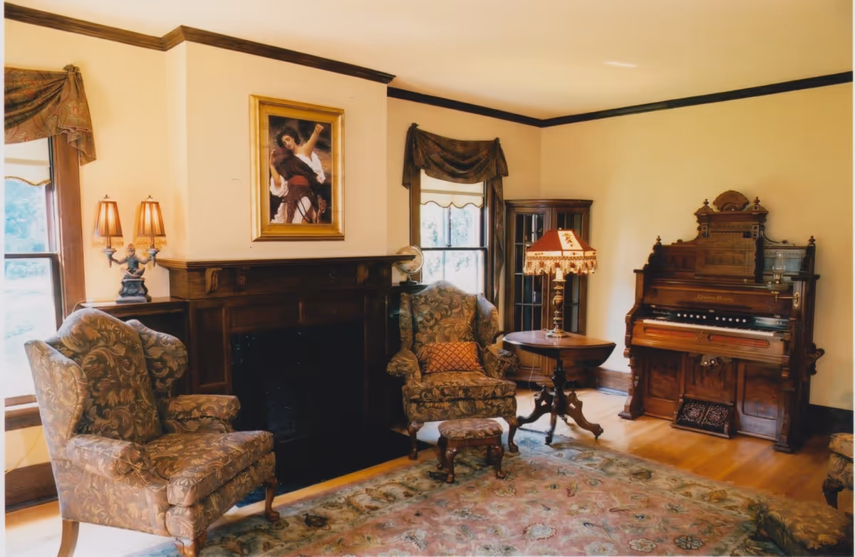 A cozy living room with two patterned armchairs, a small footstool, a wooden side table with a decorative lamp, a dark wood fireplace with a framed painting above it, and an ornate wooden organ against the wall. The room has light-colored walls with dark wood trim and two windows with draped curtains.