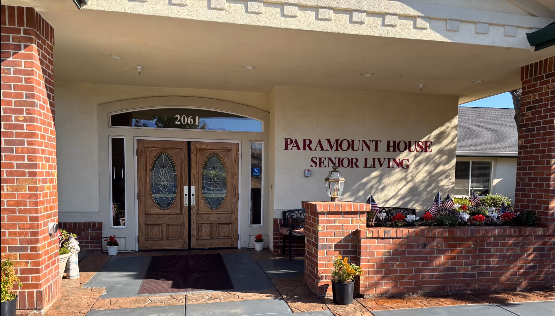 Entrance to Paramount House Senior Living facility with double wooden doors featuring oval glass panels, brick pillars, and a flower bed decorated with small American flags.