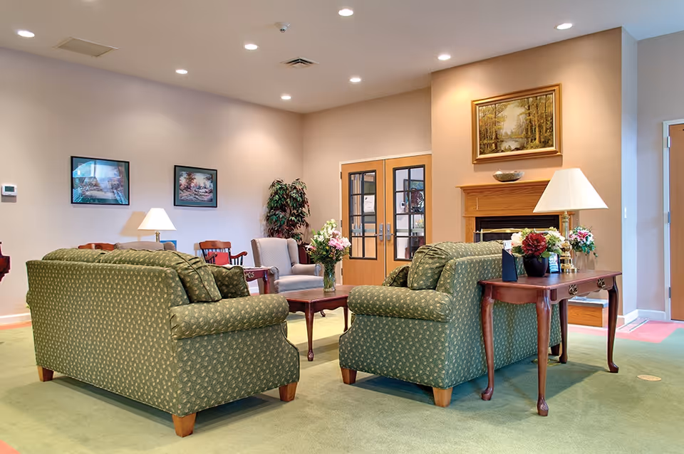 A cozy living room area with two green patterned sofas facing each other, a wooden coffee table with a vase of flowers, a side table with a lamp and flowers, a fireplace with a framed painting above it, and double doors with glass panels in the background.