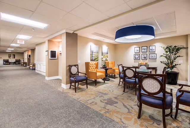 A well-lit interior common area with a seating arrangement including wooden chairs with blue cushions, two orange tufted armchairs, a small round table with flowers, and a dining table with chairs. The walls are decorated with framed pictures and there is a large potted plant in the corner. The ceiling has recessed lighting and a large circular hanging light fixture. The carpet has a patterned design and the hallway extends to the left with additional seating visible in the distance.