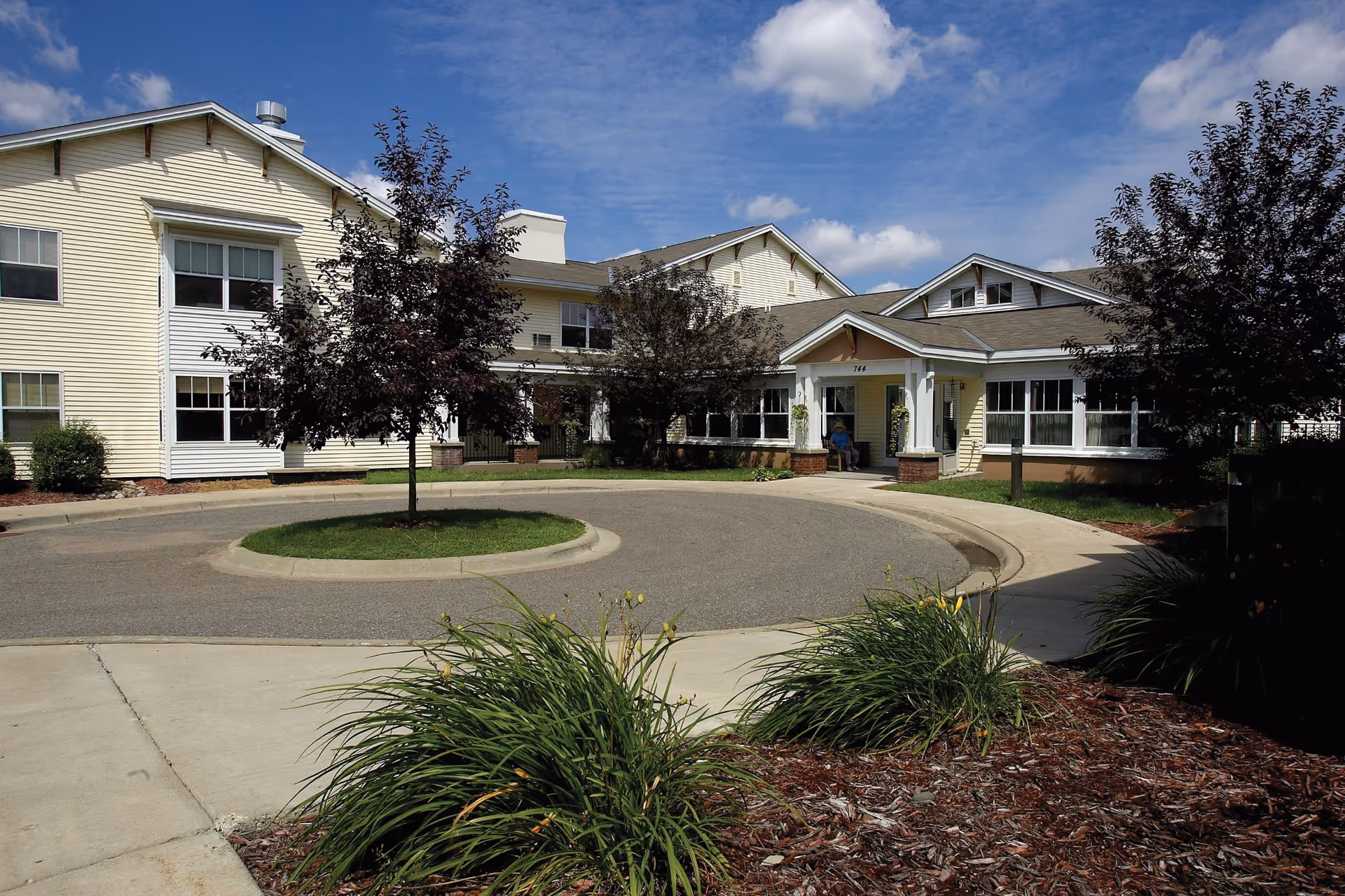 Exterior view of a senior living facility named River Heights by Vivie, showing a circular driveway with a small grassy island in the center, surrounded by a two-story yellow building with white trim and multiple windows. There are trees and landscaped bushes around the driveway, and a person sitting near the entrance under a covered porch.
