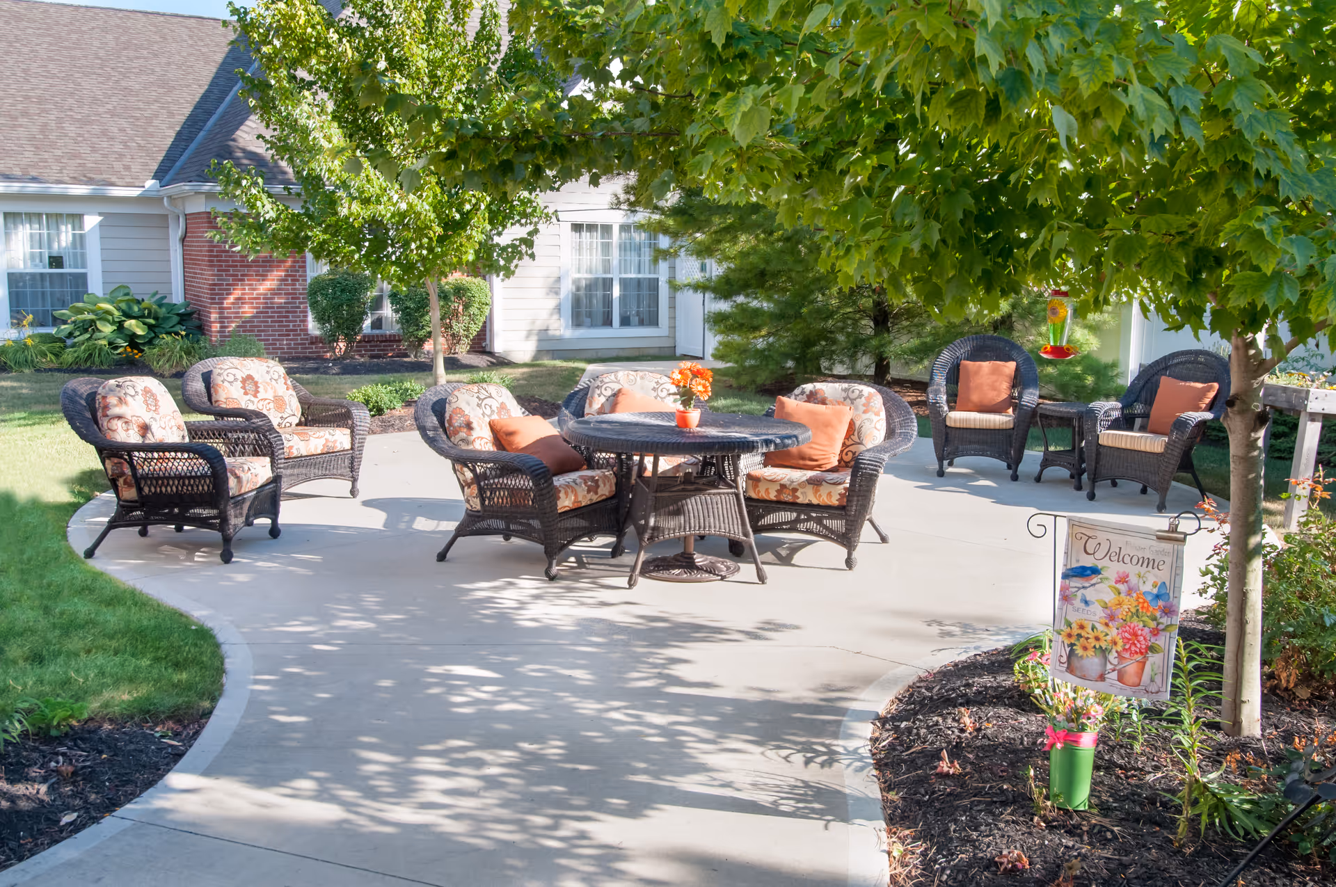 Outdoor patio with wicker seating and a round table, cushions, a 'Welcome' sign, and landscaping in front of a residential building.