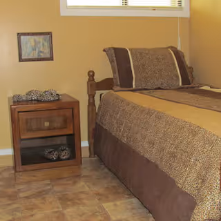 A bedroom corner featuring a bed with brown and beige bedding, a wooden nightstand with decorative items, a framed picture on the beige wall, and a window with blinds above the bed.