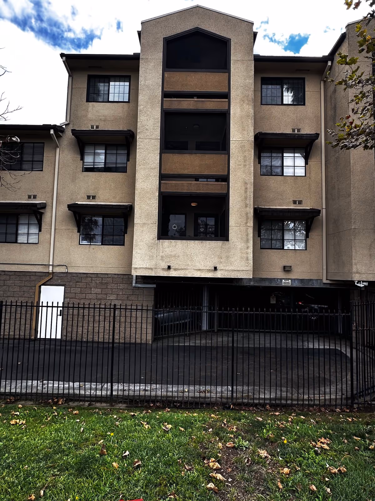 Front facade of a multi-story beige residential building with recessed balconies and a gated ground-floor parking entrance.