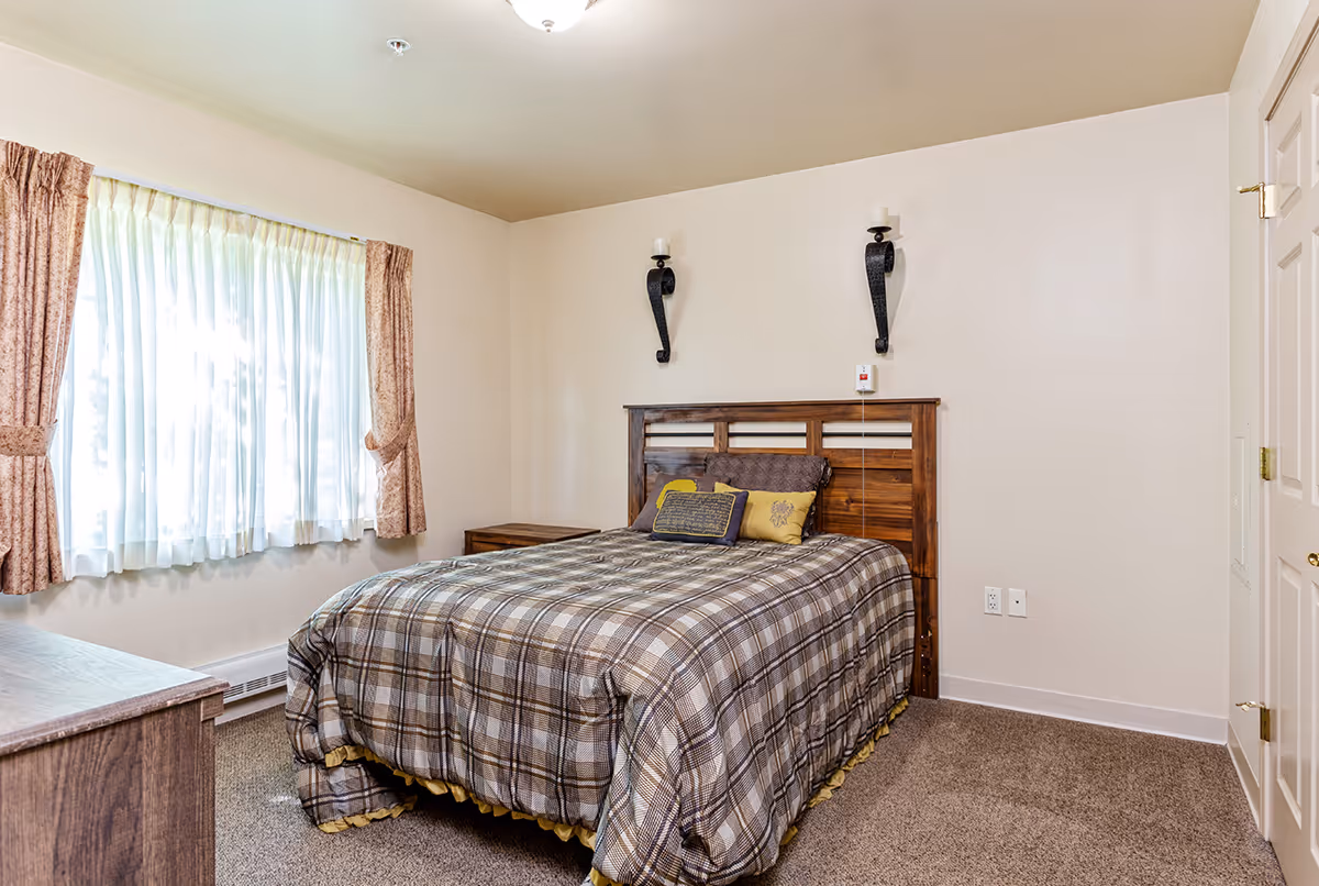 Simple furnished bedroom with a double bed, plaid comforter, wooden headboard, nightstand, curtained window, and neutral walls.