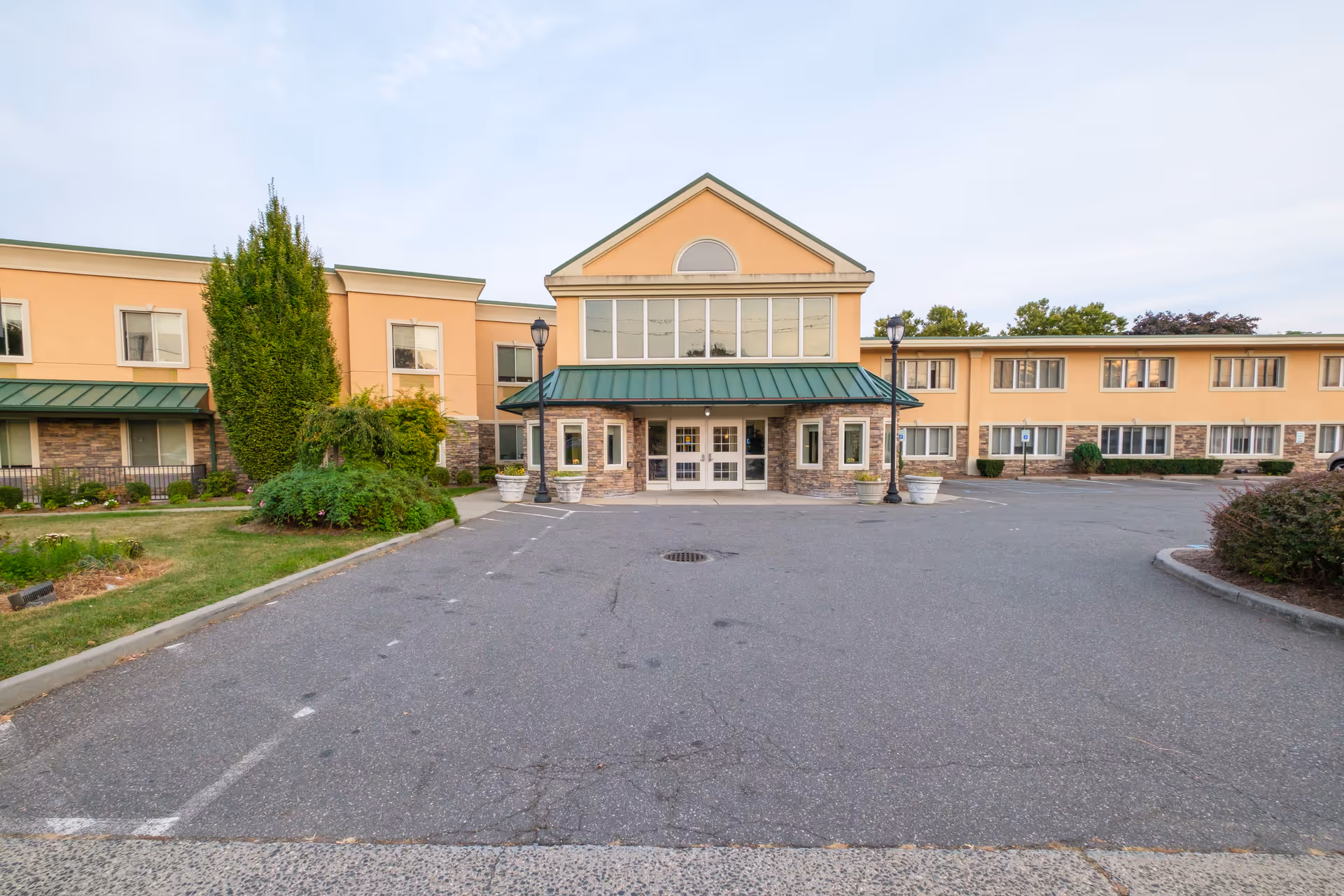 Front exterior view of Amber Court of Westbury, a two-story building with a green metal roof over the entrance, beige walls, and stone accents. There are two lamp posts and several large planters near the entrance, with a parking area in front and some greenery on the sides.