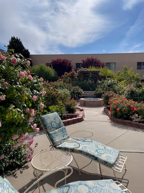 Outdoor garden area with blooming pink and red flowers, green shrubs, and a circular water fountain in the center. Two cushioned lounge chairs with a small round table are placed on a paved pathway. A building is visible in the background under a partly cloudy sky.