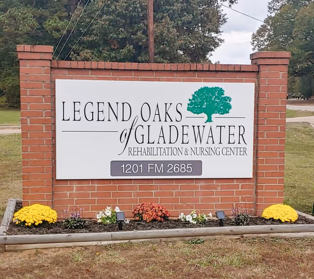 A brick sign for Legend Oaks of Gladewater Rehabilitation & Nursing Center with the address 1201 FM 2685. The sign features a green tree logo and is surrounded by a small flower bed with yellow, red, and white flowers.