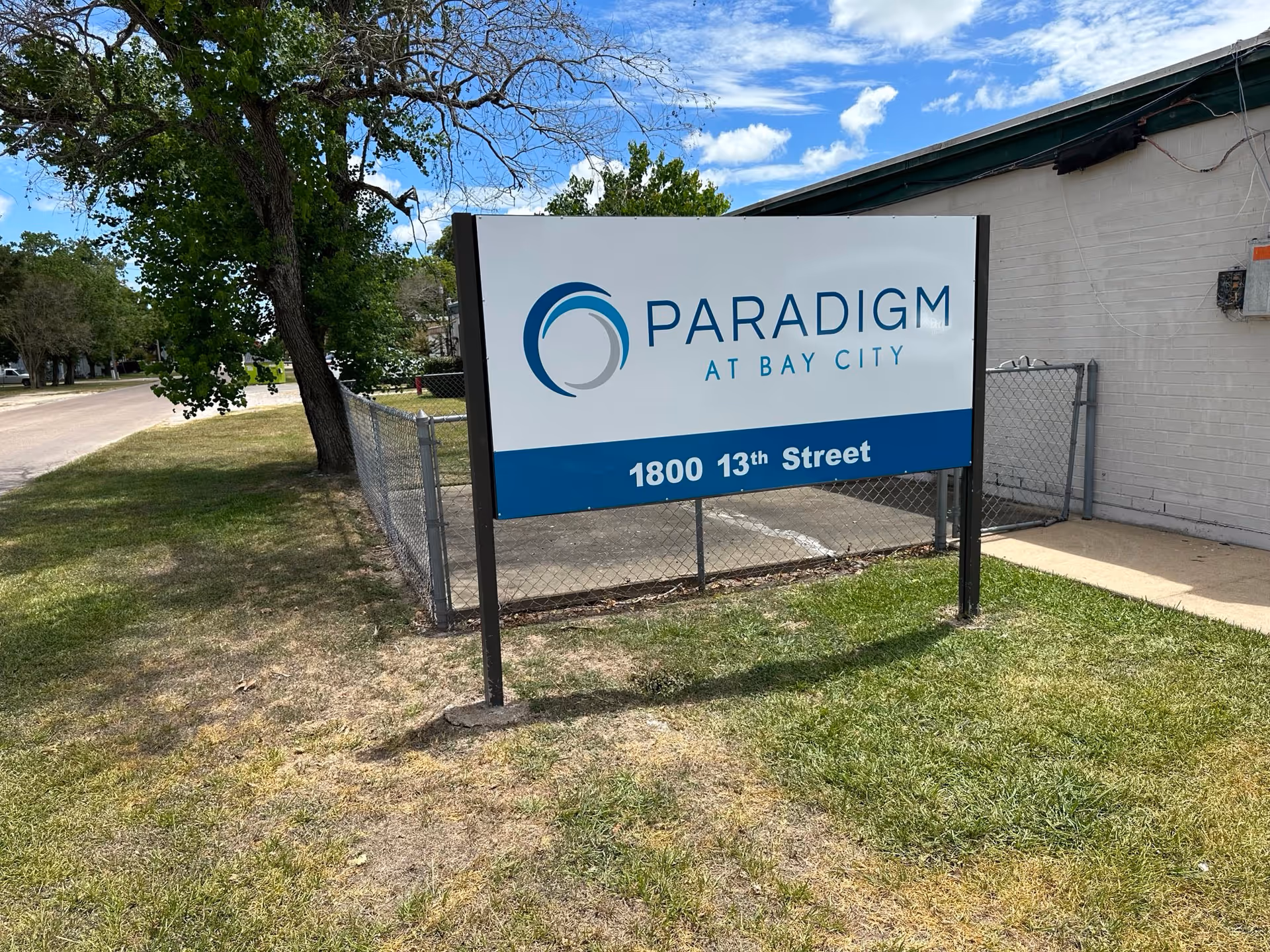 Freestanding outdoor sign reading "Paradigm at Bay City" and "1800 13th Street" in front of a building and chain-link fence.