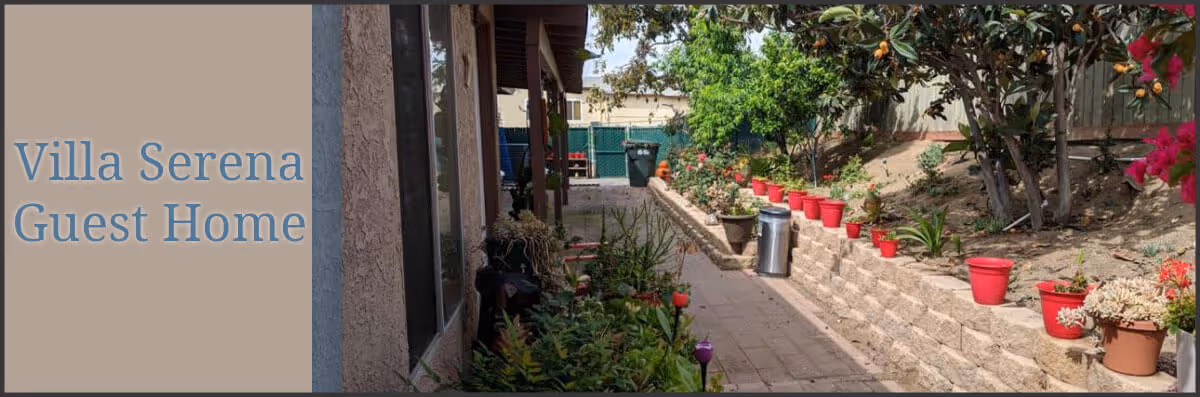 Outdoor garden area at Villa Serena Guest Home featuring a paved walkway bordered by a retaining wall with multiple red flower pots and various plants and shrubs on a sunny day.