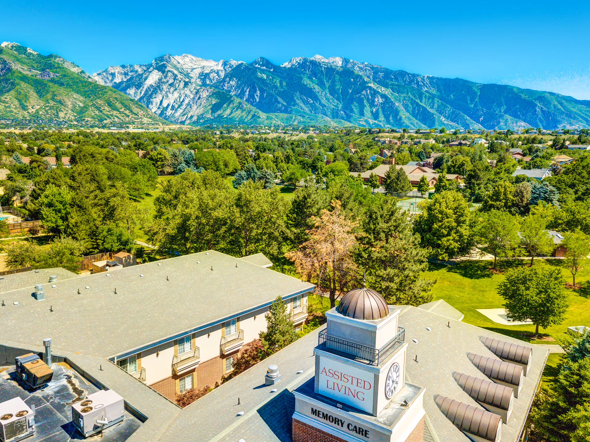 Aerial view of an assisted living and memory care building surrounded by green trees, lawns and residential areas with mountains in the background.