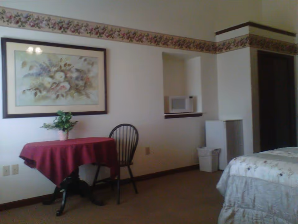 A room with a bed partially visible on the right, a small table covered with a red tablecloth and a potted plant on top, a single black chair, a floral painting on the wall, a microwave placed in a recessed shelf, a small white refrigerator, and a trash bin. The walls have a floral border near the ceiling.