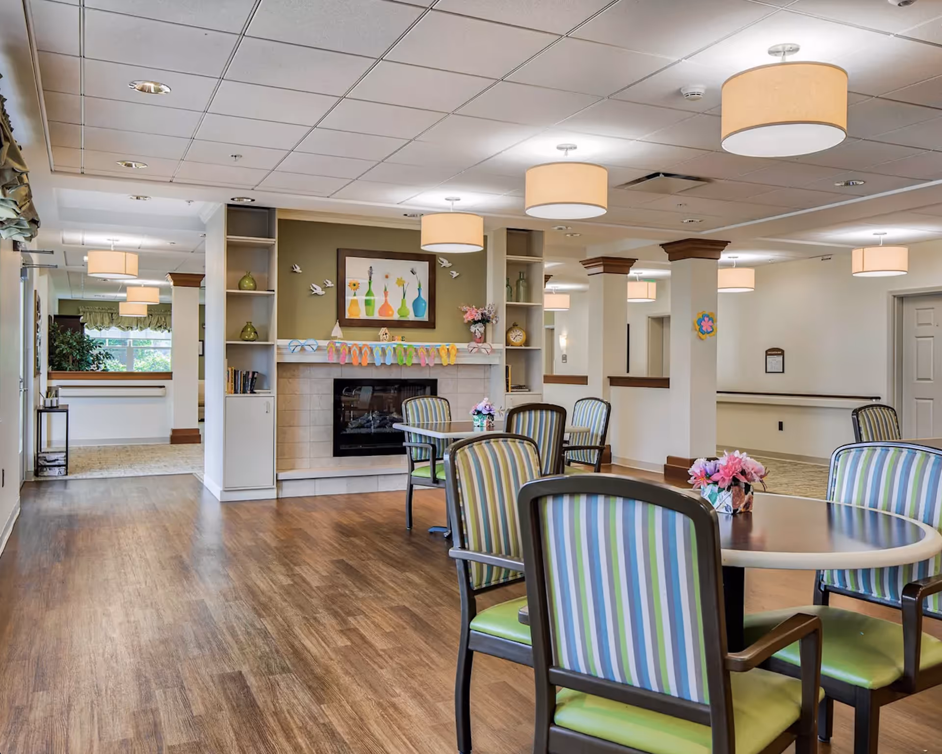 A bright and spacious common area in a senior living facility featuring a fireplace with decorative vases and a colorful garland above it. There are several round tables surrounded by striped chairs with green cushions. The room has wooden flooring, multiple ceiling lights with beige lampshades, and built-in shelves with decorative items. The walls are painted in neutral tones, and there is a hallway visible in the background.