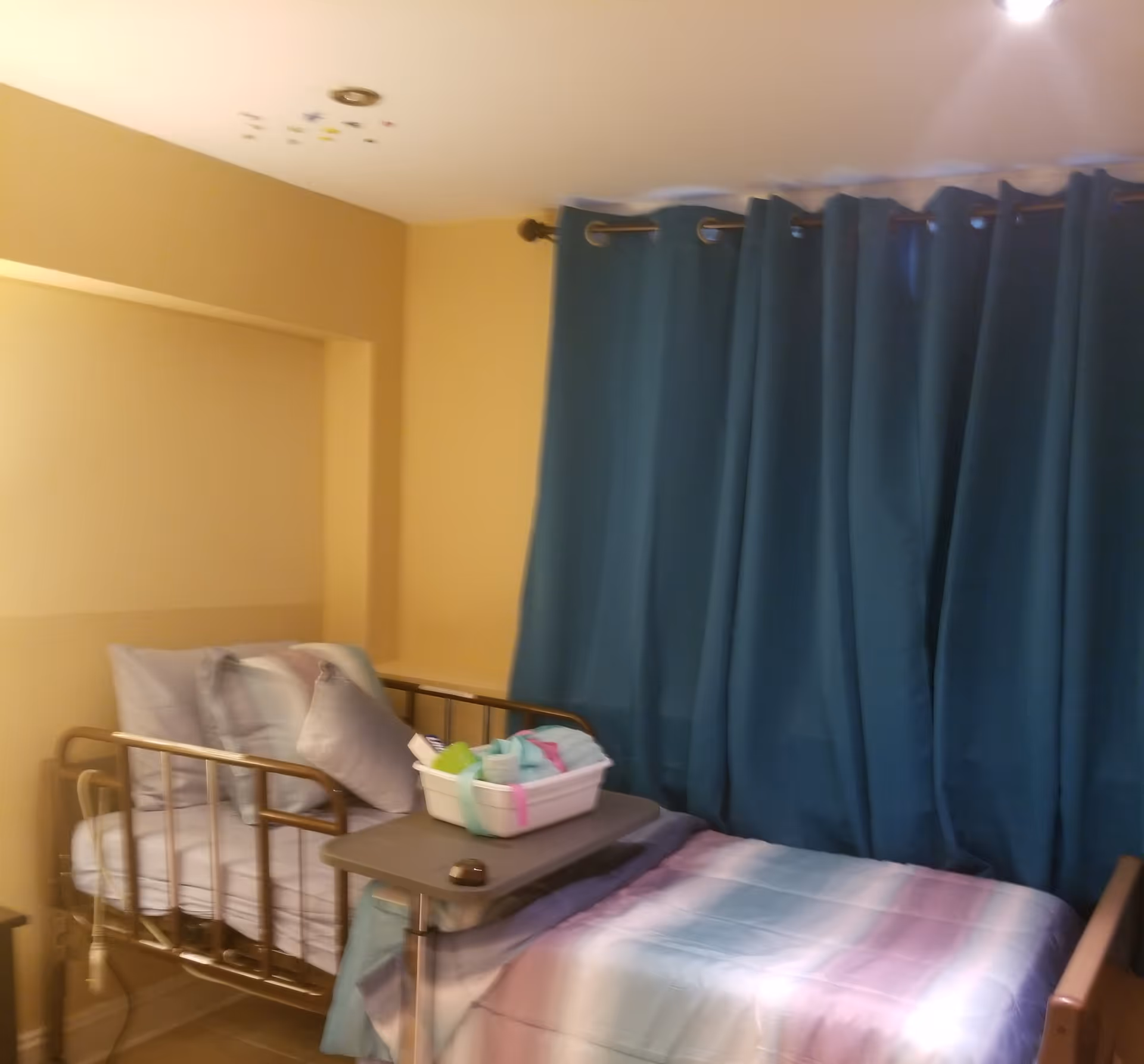 A small bedroom with a single bed featuring metal rails and multiple pillows. The bed is covered with a pastel-colored blanket. Next to the bed is a small overbed table holding a white basket with various items. The room has beige walls and a large dark blue curtain covering the window.