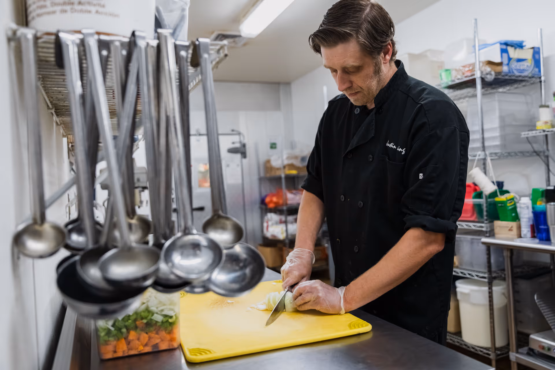 A chef wearing a black uniform and gloves is chopping an onion on a yellow cutting board in a commercial kitchen. Several metal ladles hang on a rack in the foreground, and shelves with various kitchen supplies are visible in the background.