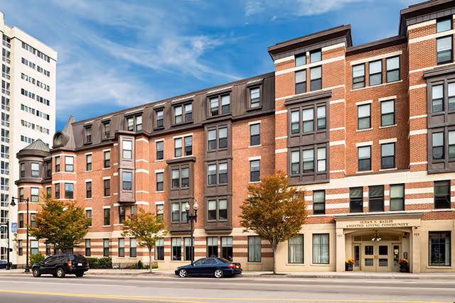 Exterior view of a multi-story brick building with large windows and a sign above the entrance that reads 'Susan S Bailis Assisted Living Community'. There are a few trees and parked cars along the street in front of the building under a partly cloudy blue sky.
