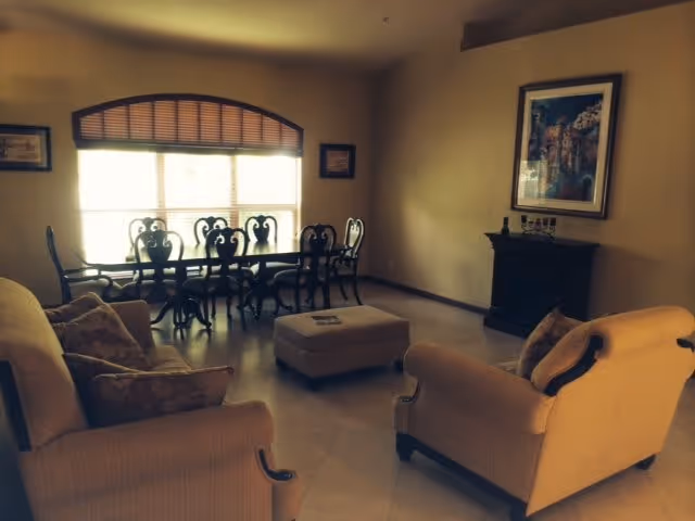A cozy living room with two beige armchairs and a matching sofa arranged around a central ottoman. In the background, there is a dining table with six ornate chairs positioned in front of a large window with wooden blinds. A framed painting hangs on the wall above a small black fireplace or cabinet.
