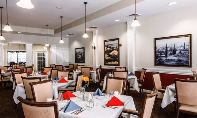A dining room with multiple tables covered in white tablecloths, each set with glasses, cups, silverware, and folded red and blue napkins. The room has beige walls with framed landscape paintings and pendant lights hanging from the ceiling.