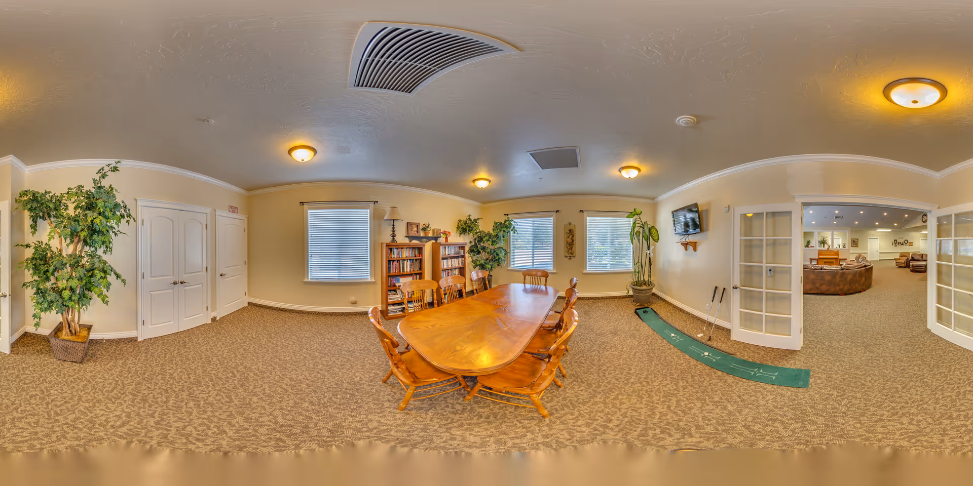 A spacious room with a long wooden table surrounded by wooden chairs, two large windows with blinds, bookshelves filled with books and decorative items, potted plants, a wall-mounted TV, and a putting green mat with golf clubs. The room has beige walls, carpeted floor, and ceiling lights. French doors open to another room with seating areas.