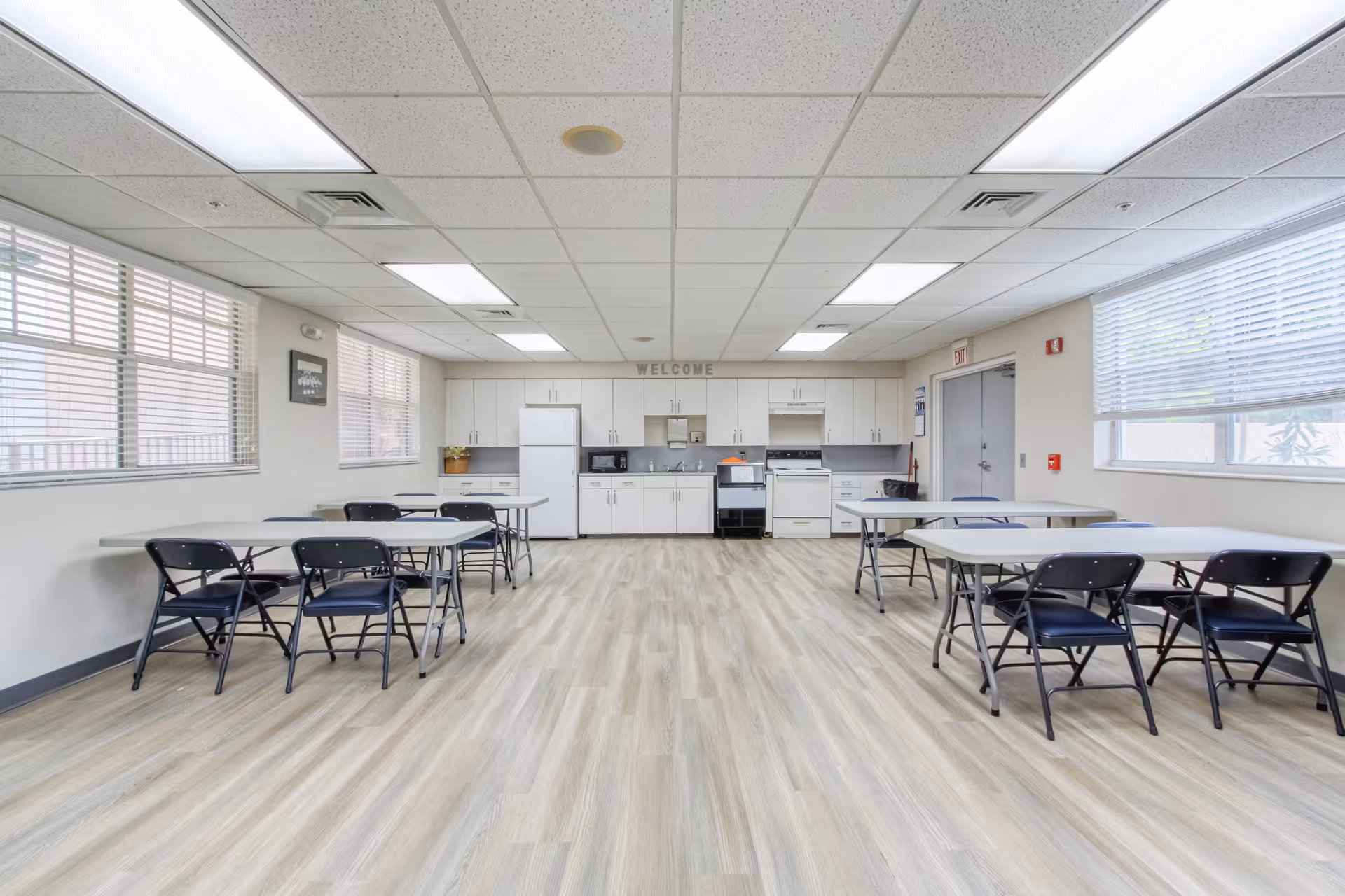 A bright and clean communal kitchen and dining area with white cabinets, a refrigerator, microwave, stove, and multiple folding tables with black folding chairs. Large windows with blinds allow natural light to fill the room, and a 'WELCOME' sign is displayed above the cabinets.