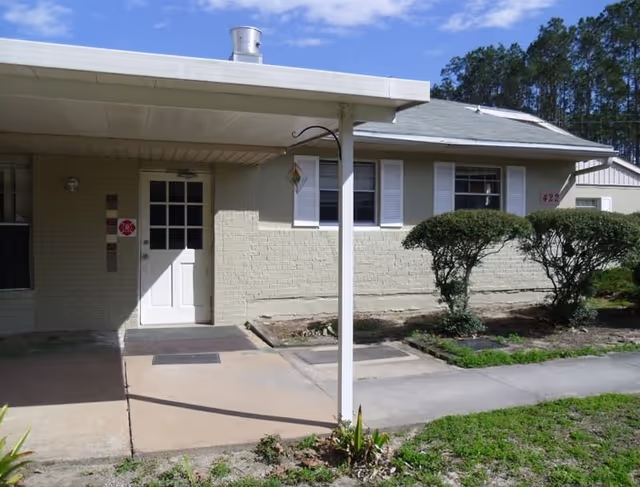 Exterior view of a single-story building with light-colored brick walls, white door with glass panes, and white window shutters. There is a covered entrance supported by a white post, some trimmed bushes, and a concrete walkway leading to the door. The sky is clear with a few clouds.