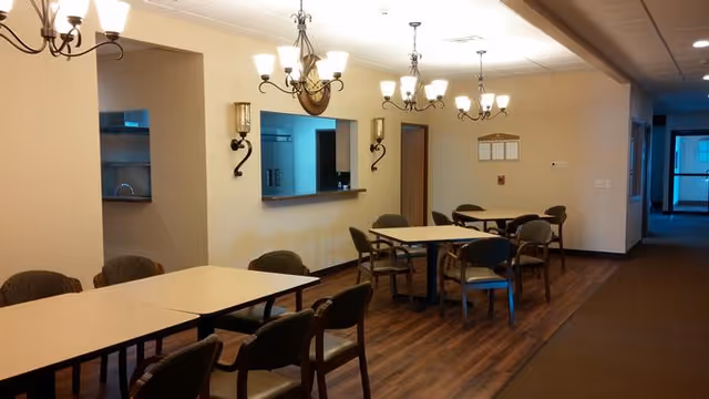 Interior view of a dining area in a senior living facility with multiple tables and chairs arranged on a wooden floor. The room is lit by chandeliers and wall sconces, with a serving window and door visible on the far wall.