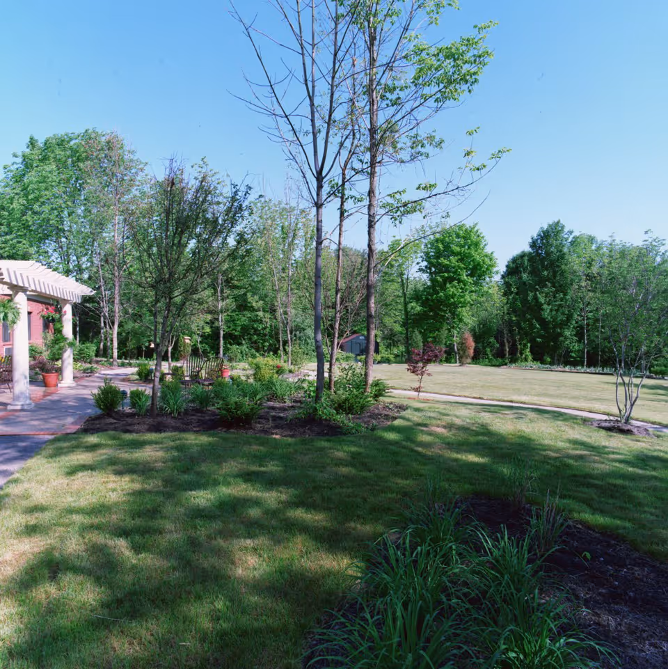 A landscaped outdoor garden area with green grass, various trees, shrubs, and plants under a clear blue sky. A paved walkway leads to a building with a white pergola on the left side of the image.