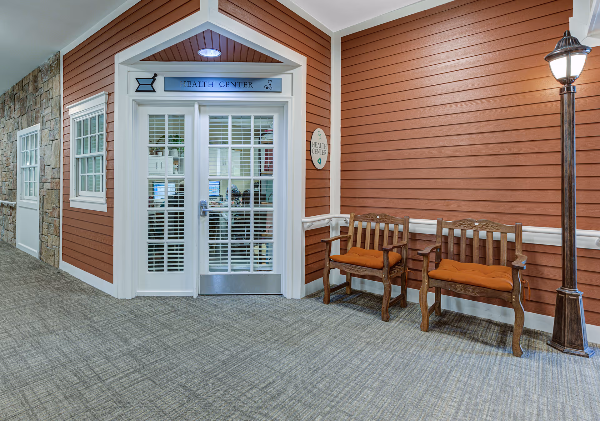 Hallway leading to a Health Center with white-framed glass double doors, two wooden chairs with orange cushions against a red paneled wall, and a standing lamp.