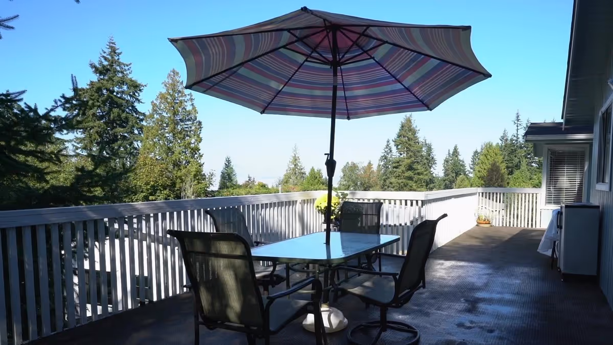 Outdoor patio area with a glass-top table and four black mesh chairs arranged around it. A large striped umbrella is open above the table providing shade. The patio is surrounded by a white wooden railing and overlooks a view of tall green trees under a clear blue sky. There are some potted plants near the railing and the side of the building.