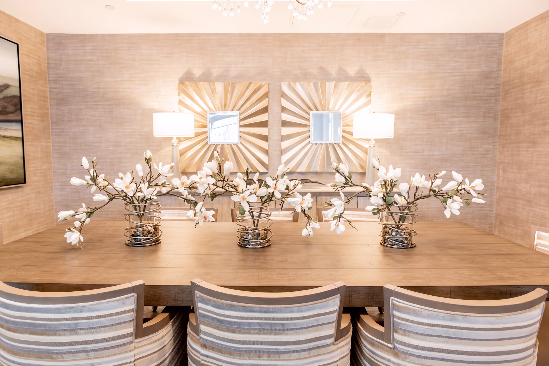 A dining room with a wooden table decorated with three vases containing white flowers. Four striped upholstered chairs are positioned around the table. The background features two decorative sunburst mirrors and two table lamps with white shades on a console table against a textured beige wall.