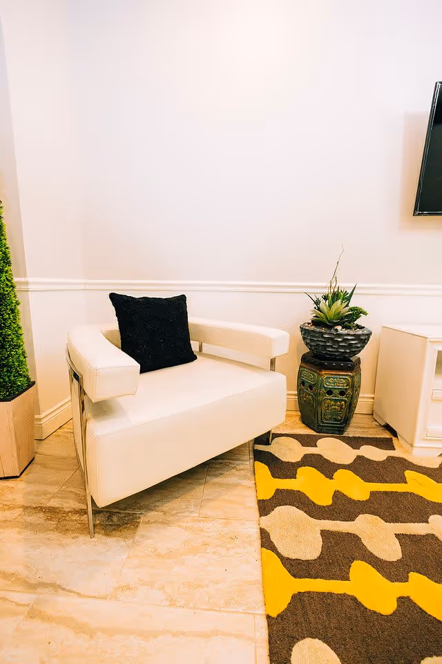 A modern white armchair with a black cushion is placed on a tiled floor next to a colorful rug with yellow, brown, and beige patterns. To the right of the chair is a decorative green ceramic plant stand with a potted plant on top. The wall behind is plain white with a white chair rail molding.