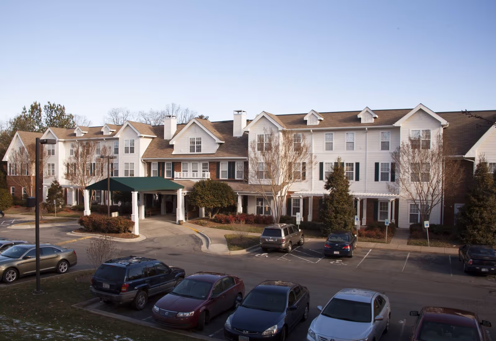 Exterior view of a multi-story senior living facility building with a covered entrance and several parked cars in front. The building has white siding with brick accents, multiple windows, and a pitched roof with dormer windows. Trees and shrubs are planted around the entrance and parking area under a clear sky.
