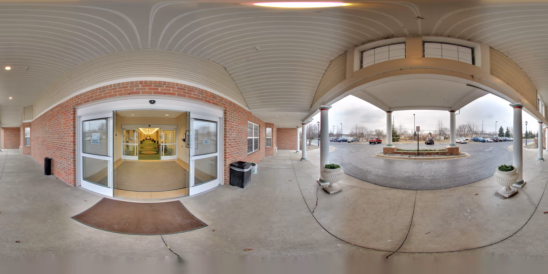 Covered entrance area of a building with automatic sliding glass doors leading inside. The exterior walls are made of red brick. There are two large white planters on either side of the entrance and a circular driveway with parked cars visible in the background under an overcast sky.