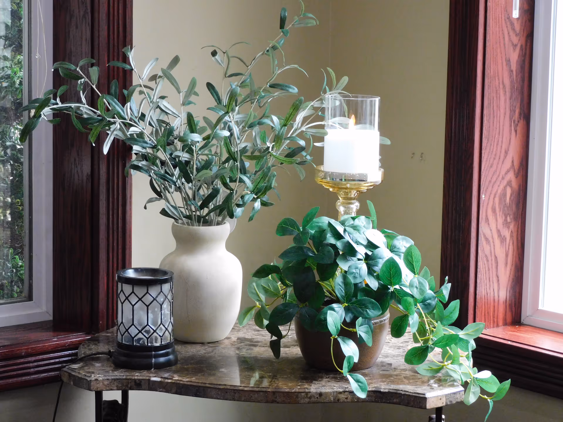 A marble-top table positioned between two windows with dark wooden frames, holding a white vase with green leafy branches, a small black lantern, a potted plant with green leaves, and a glass candle holder with a lit white candle.