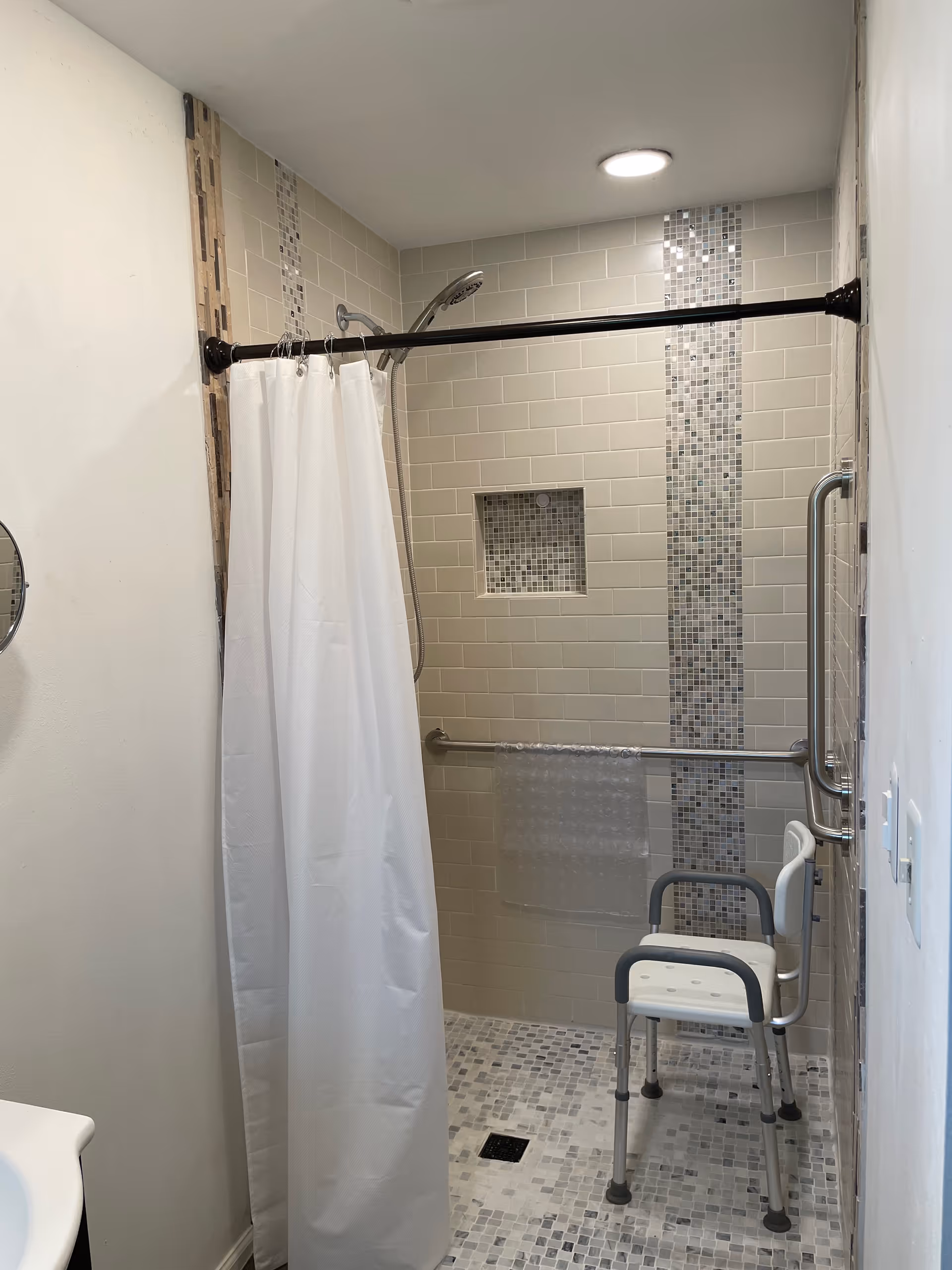 Accessible tiled shower with a white curtain, shower chair, grab bars, recessed niche and mosaic tile accents.