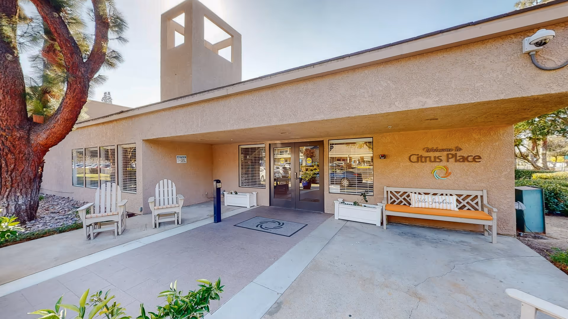 Entrance to Citrus Place facility with a covered walkway, two Adirondack chairs on the left, a bench with an orange cushion and a 'Hello Sunshine' pillow on the right, and a large tree to the left side. The building has beige stucco walls and a sign that reads 'Welcome to Citrus Place' near the bench.