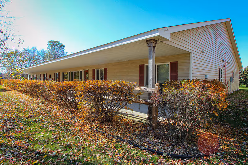Exterior view of a single-story building with beige siding and red shutters, surrounded by bushes with autumn leaves and a grassy area covered with fallen leaves under a clear blue sky.